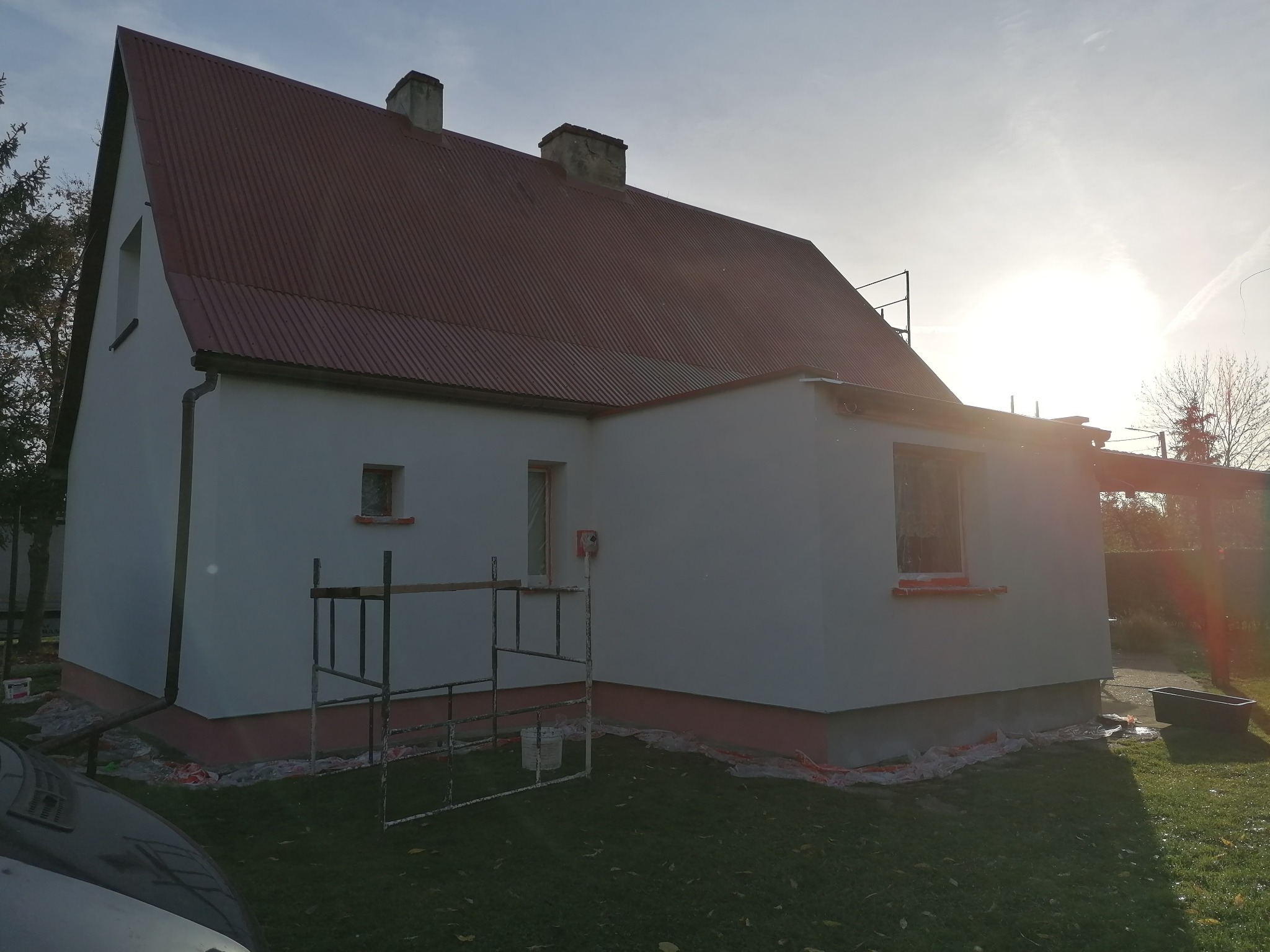 Dom jednorodzinny z czerwoną blachodachówką po tynkowaniu elewacji, widoczne rusztowanie i zabezpieczenia okien folią malarską.