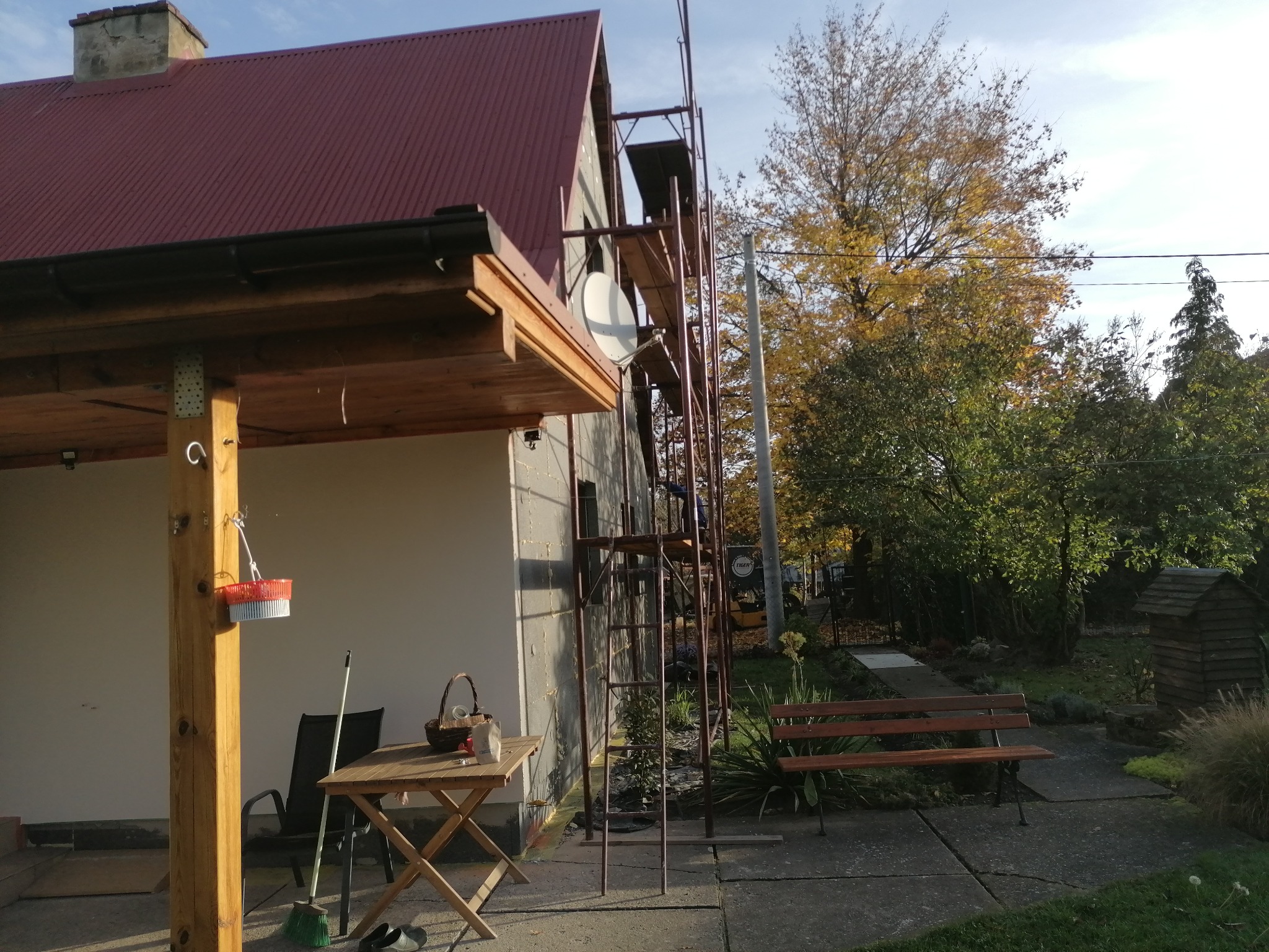 Dom jednorodzinny w trakcie prac na elewacji, widoczne rusztowanie, ocieplenie styropianem i siatka z klejem, w tle jesienne drzewa, na pierwszym planie drewniany stół i krzesło.