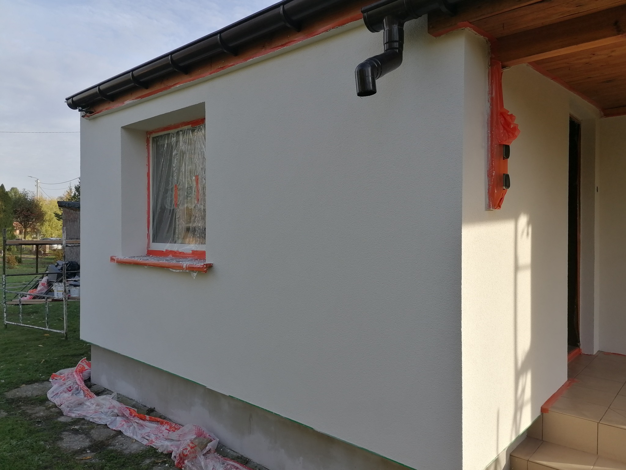 Świeżo otynkowana elewacja domu jednorodzinnego, okna i narożniki zabezpieczone folią malarską, widoczny system rynnowy i częściowo odsłonięte drewniane podbitki dachowe.
