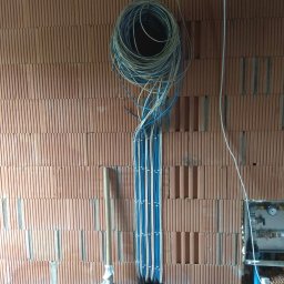 MT Systemy Marcin Taborek - Ściana z czerwonej cegły z pionowo ułożonymi kablami elektrycznymi w kolorze białym i niebieskim, zwiniętymi w pętlę u góry; poniżej widoczne czarne karbowane rury i łopata.