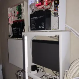 Otwarta szafka z centralą alarmową, akumulatorem podtrzymującym zasilanie i okablowaniem, widoczne diody sygnalizacyjne na płytce drukowanej i klucze do szafki.