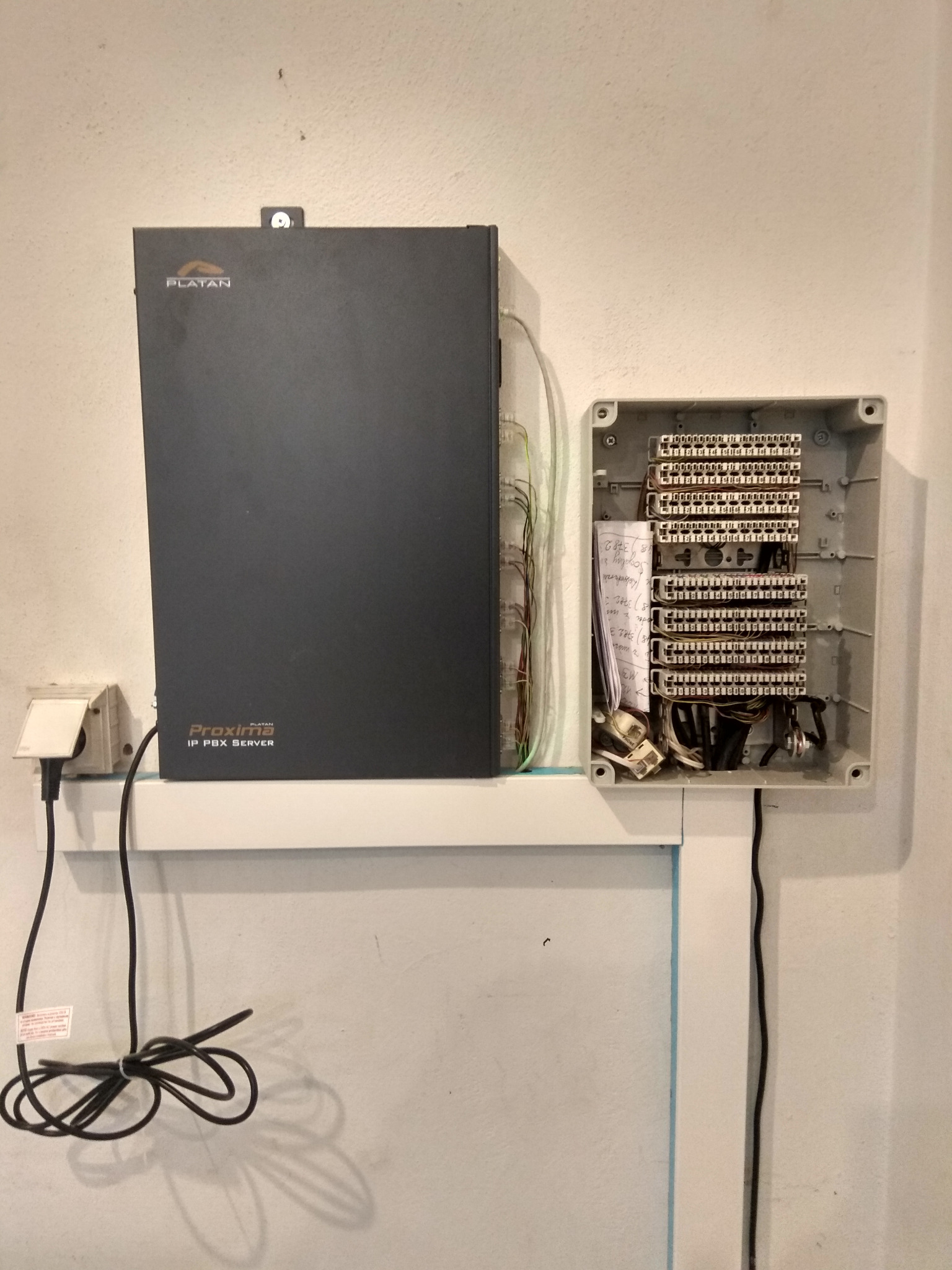 Centrala telefoniczna Platan Proxima IP PBX Server zamontowana na ścianie obok skrzynki rozdzielczej z okablowaniem.