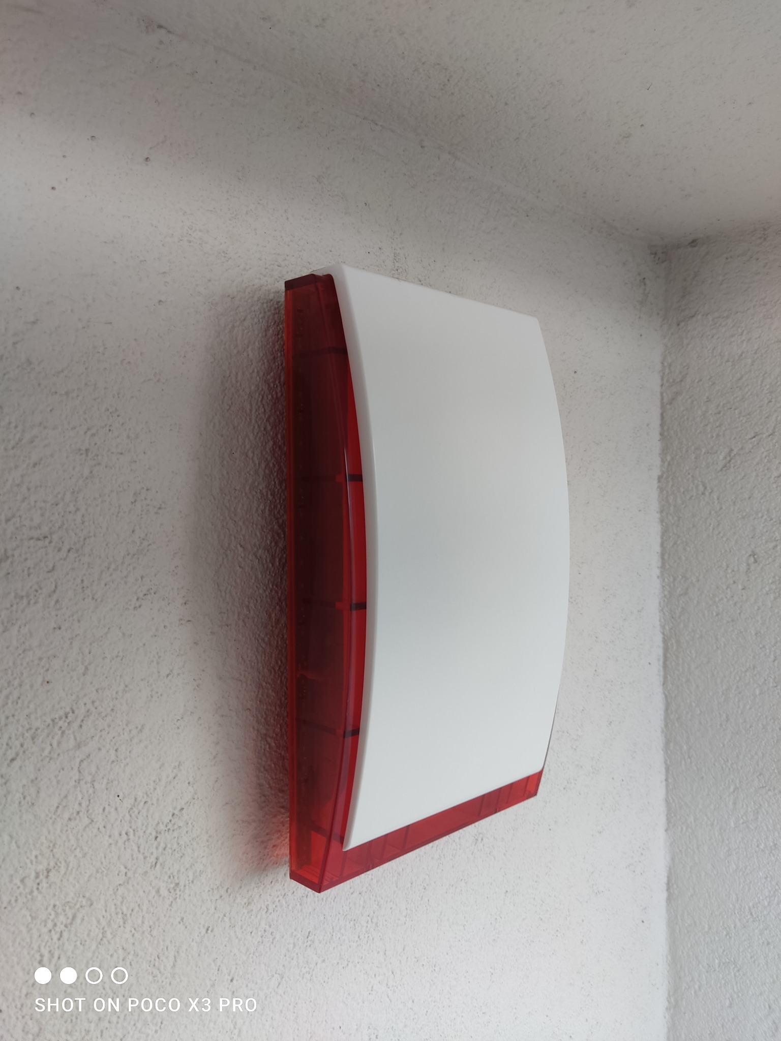 Biało-czerwona zewnętrzna syrena alarmowa zamontowana na szarej, chropowatej ścianie w rogu budynku.