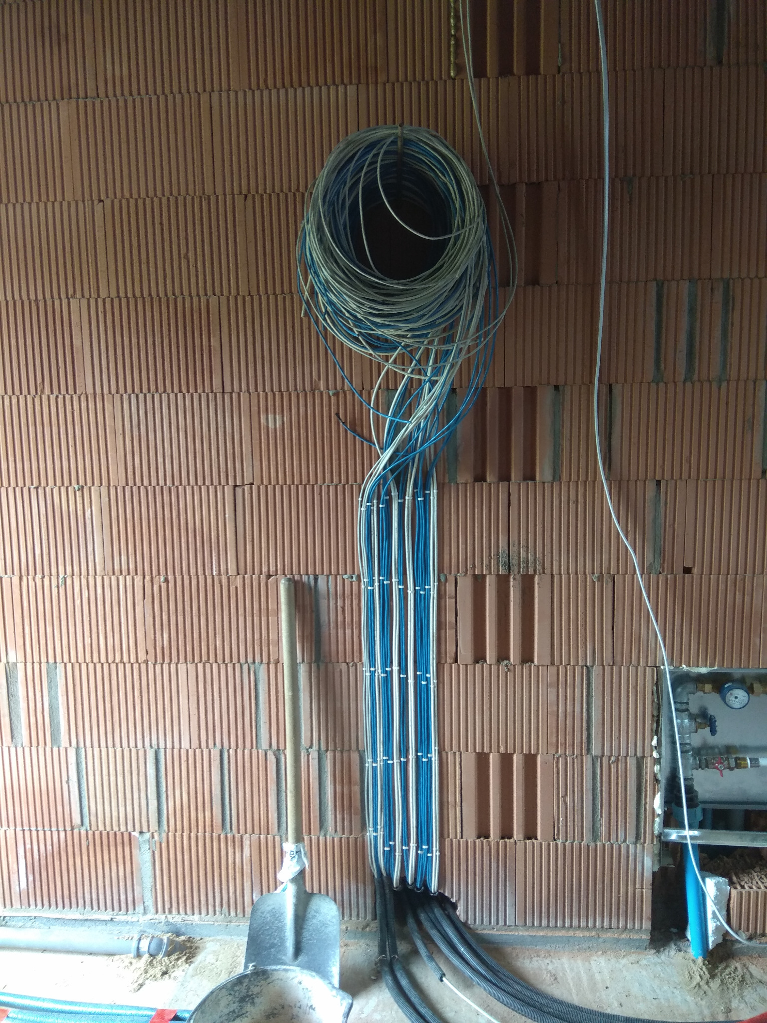 Ściana z czerwonej cegły z pionowo ułożonymi kablami elektrycznymi w kolorze białym i niebieskim, zwiniętymi w pętlę u góry; poniżej widoczne czarne karbowane rury i łopata.