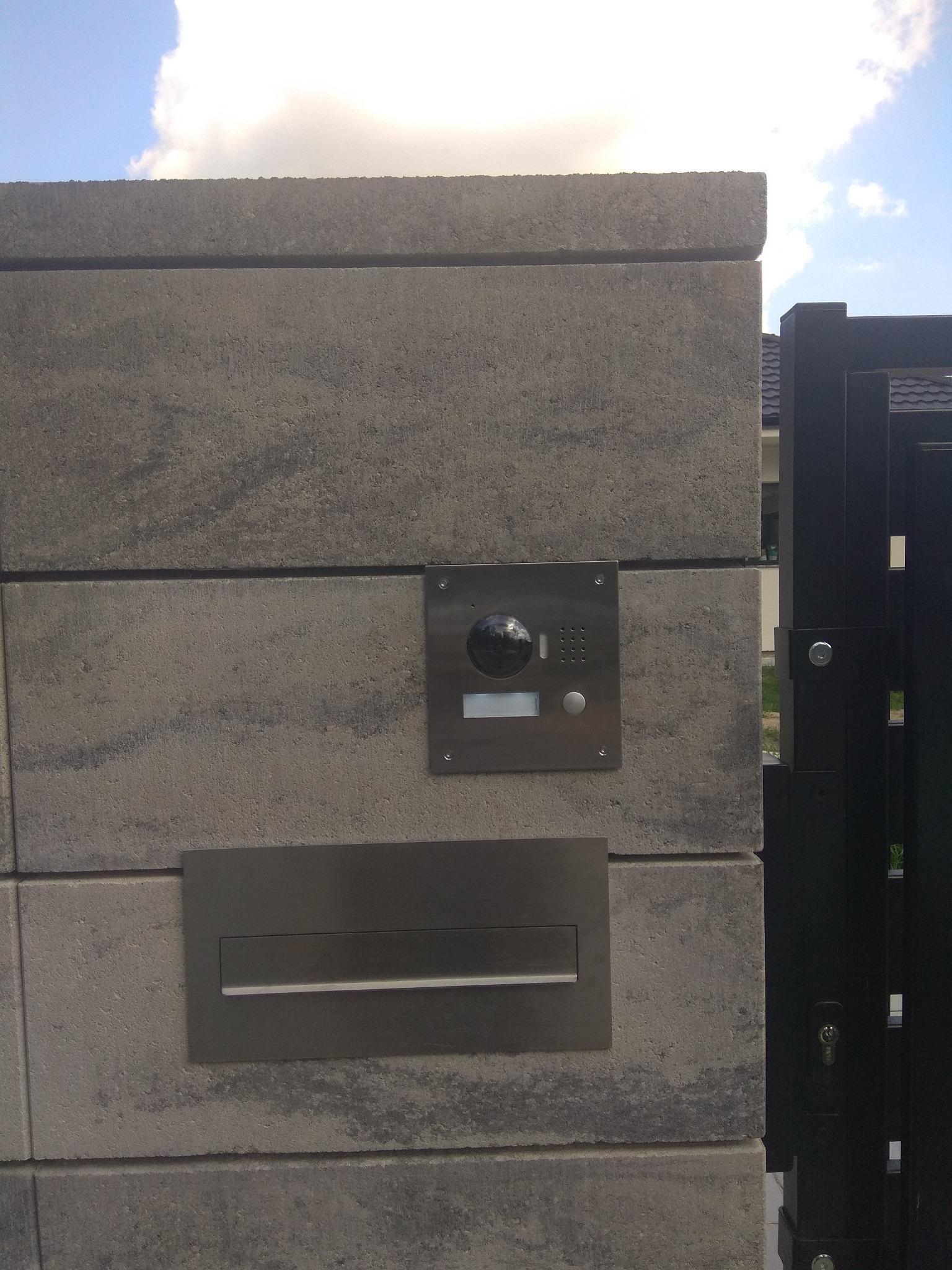 Szary, betonowy słupek ogrodzeniowy z zamontowanym wideodomofonem ze stali nierdzewnej i skrzynką na listy, widoczna czarna brama w tle oraz fragment nieba z chmurami.