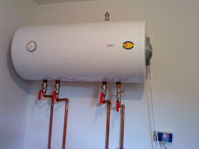 Bojler elektryczny zamontowany na ścianie w Załom, z widocznymi miedzianymi rurami i zaworami z czerwonymi uchwytami, oraz elektronicznym panelem sterowania po prawej stronie.