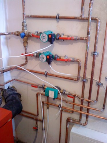 Kompleksowa instalacja grzewcza z miedzianymi rurami, pompami obiegowymi i zaworami odcinającymi, widoczne elementy kotłowni w dolnej części kadru.