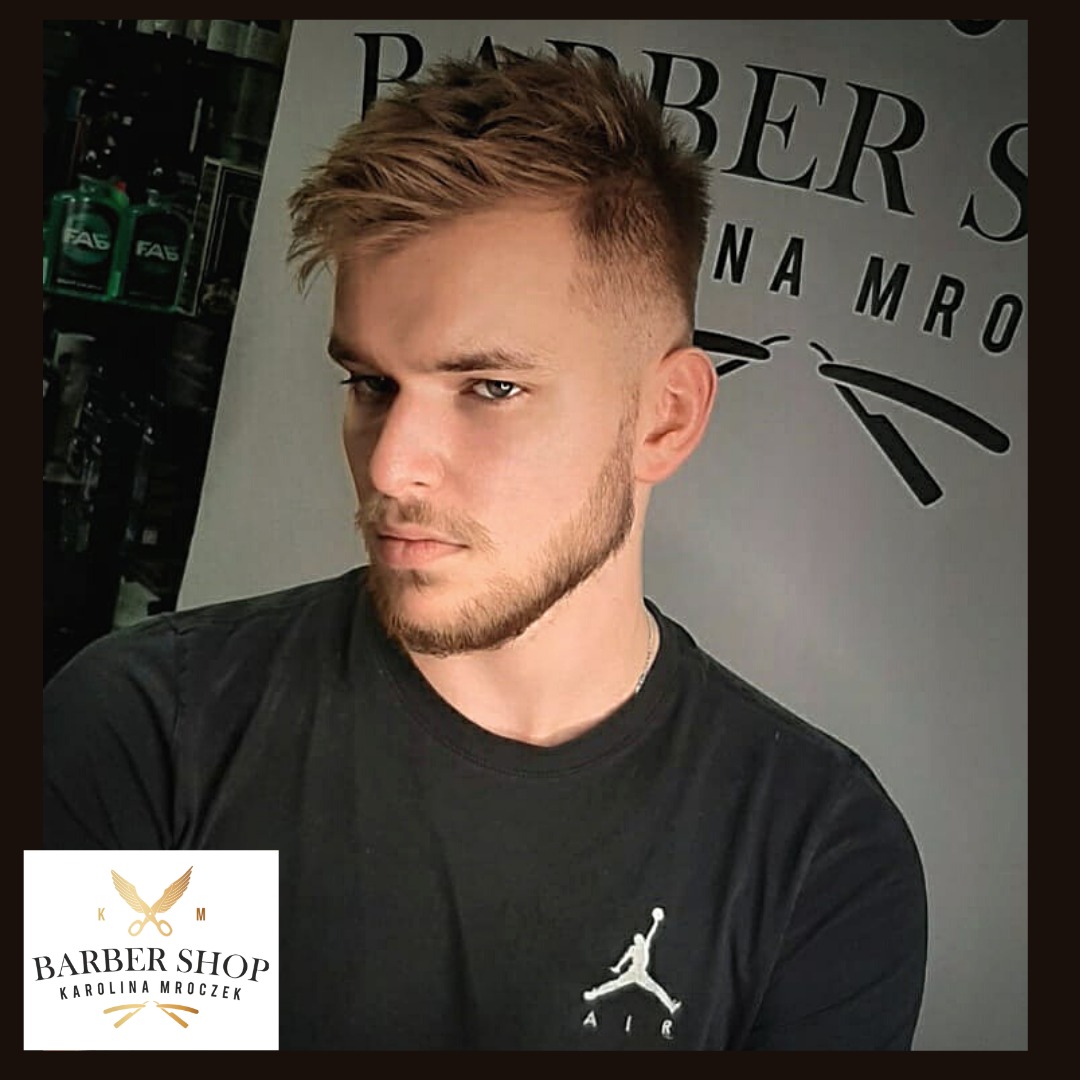Młody mężczyzna z modną fryzurą fade i krótką brodą, pozujący na tle ściany z napisem 'BARBER SHOP'.