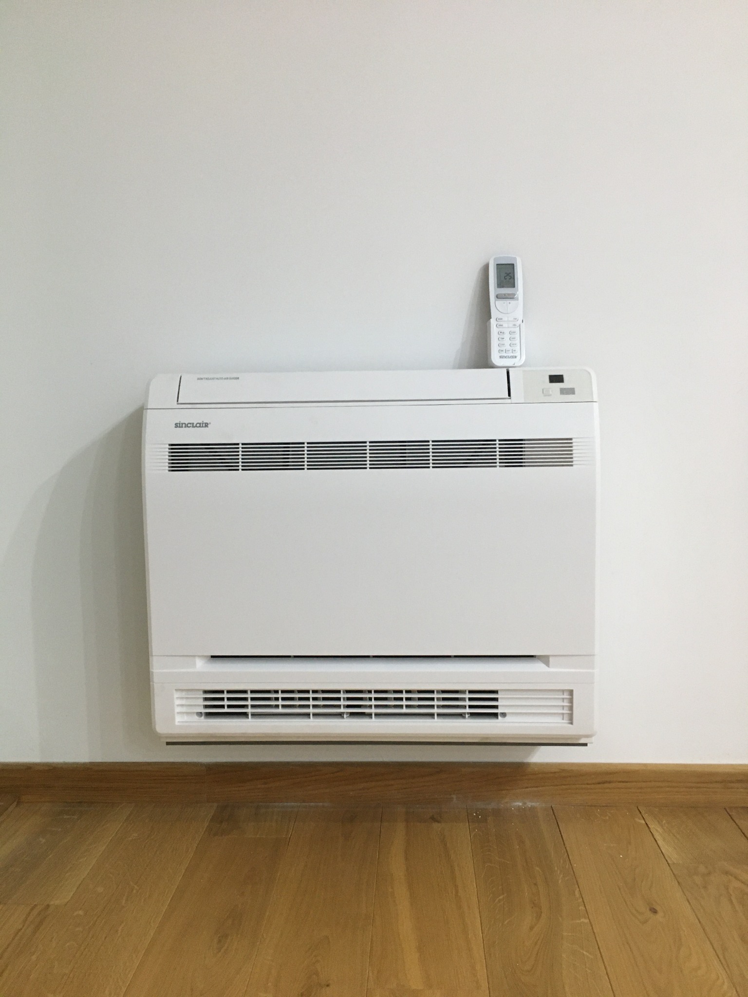 Biały klimatyzator podłogowy Sinclair z pilotem na ścianie, widok z przodu na drewnianą podłogę i listwę przypodłogową.