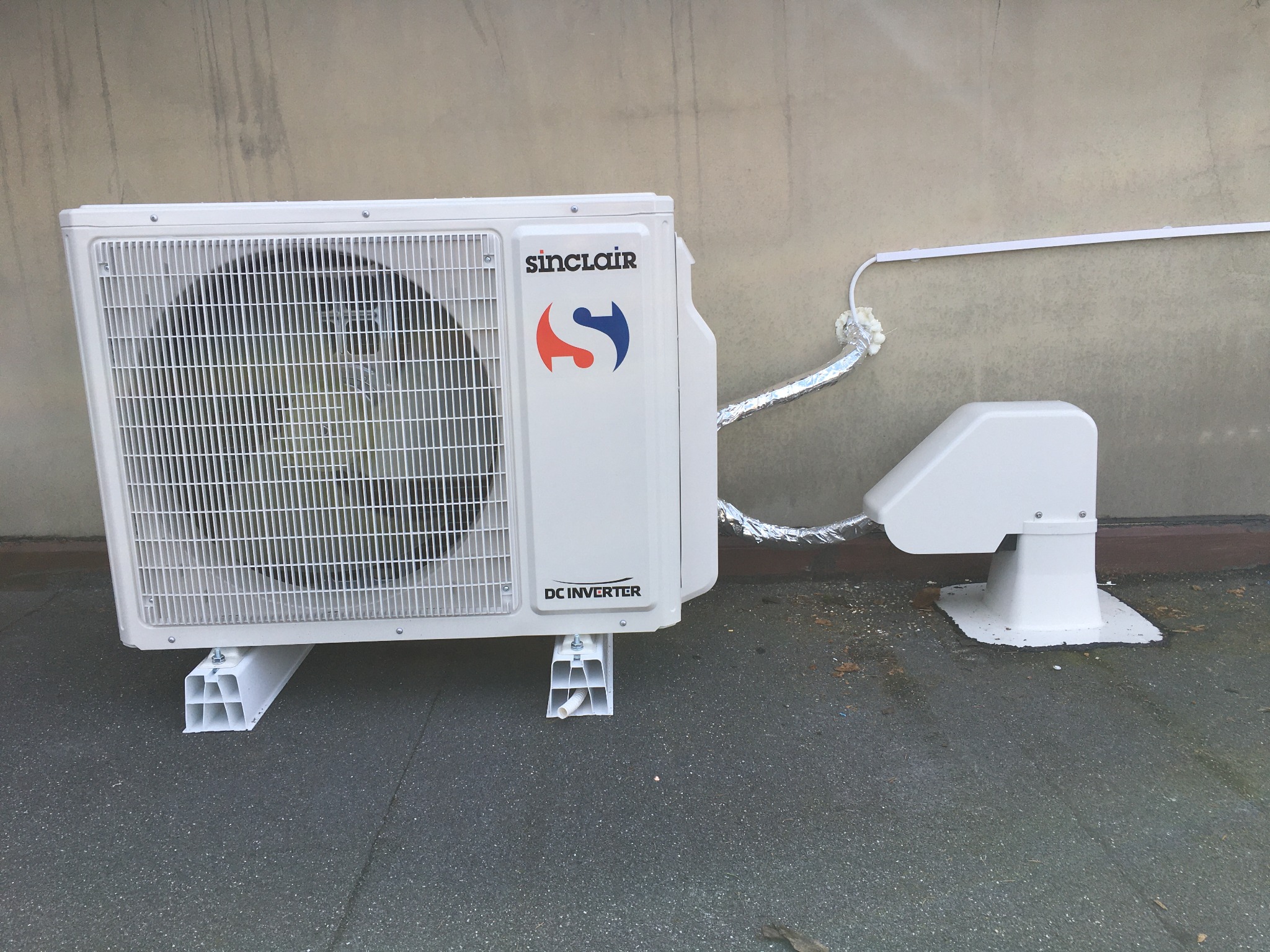 Biała jednostka zewnętrzna klimatyzacji Sinclair zamontowana na plastikowych wspornikach, z widocznymi rurami owiniętymi folią aluminiową i uszczelnieniami z pianki montażowej.