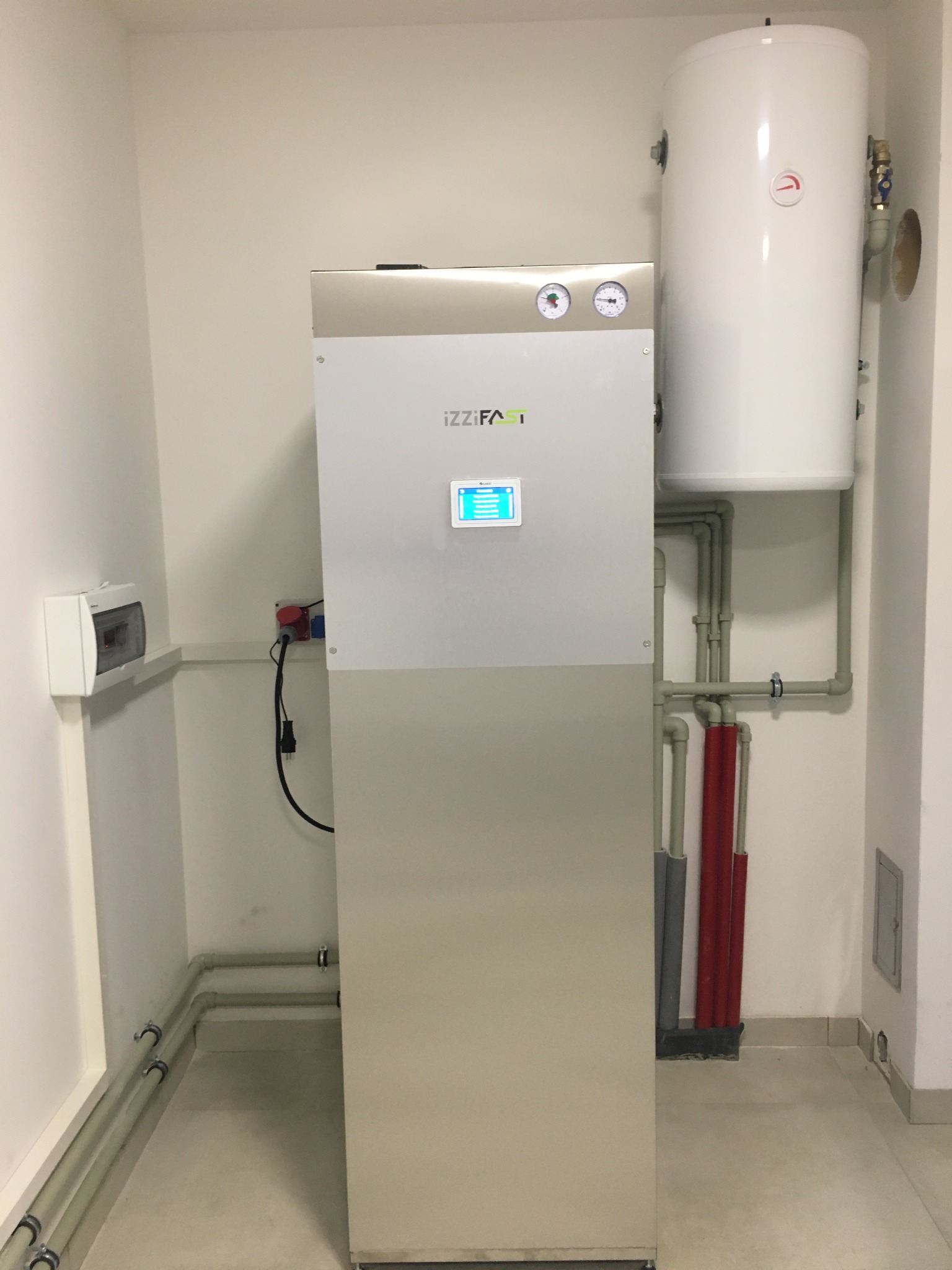 Nowoczesna pompa ciepła IZZI FAST z zasobnikiem ciepłej wody w pomieszczeniu technicznym, widoczne rury instalacyjne i skrzynka elektryczna.