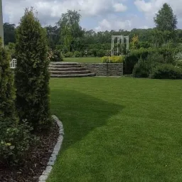 Zadbany trawnik z widocznym obrzeżem rabaty z kostki granitowej, w tle kamienne schody i biała altana ogrodowa, drzewa i krzewy.