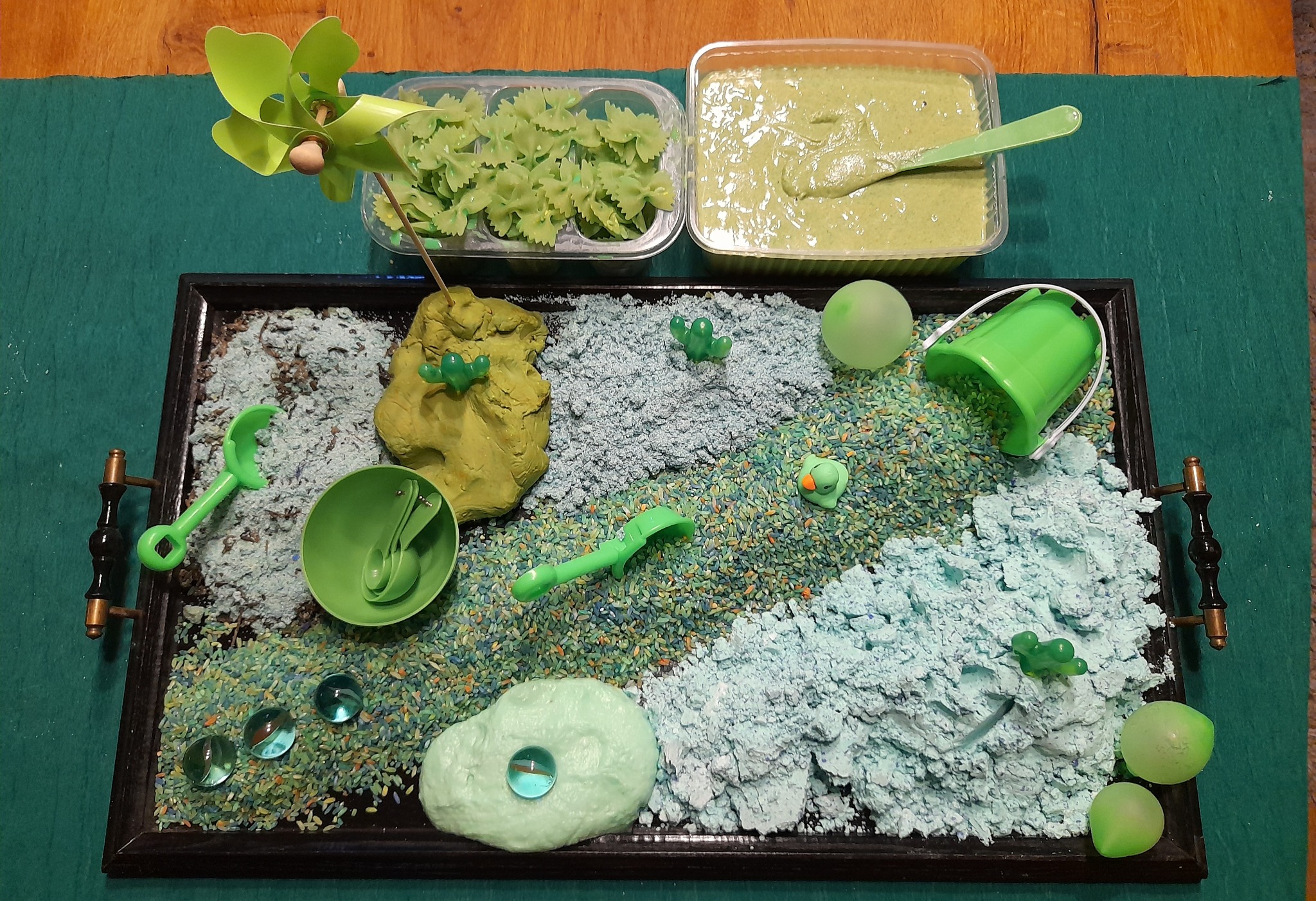 Sensoryczny stolik z zielonymi elementami: makaron motylkowy, masa plastyczna, piasek kinetyczny, zabawki i kule, całość na czarnej tacy z uchwytami.