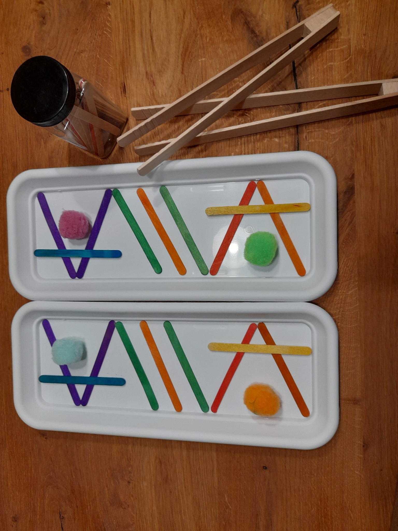 Dwa białe pojemniki z kolorowymi patyczkami i pomponami ułożonymi w geometryczne wzory, obok leżą drewniane szczypce i słoik z patyczkami na drewnianym stole.