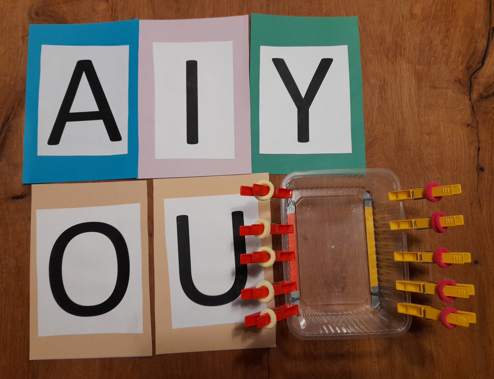 Edukacyjna pomoc: kolorowe karty z literami A, I, Y, O, U oraz pojemnik z klamerkami z gąbkami do nauki przez zabawę.