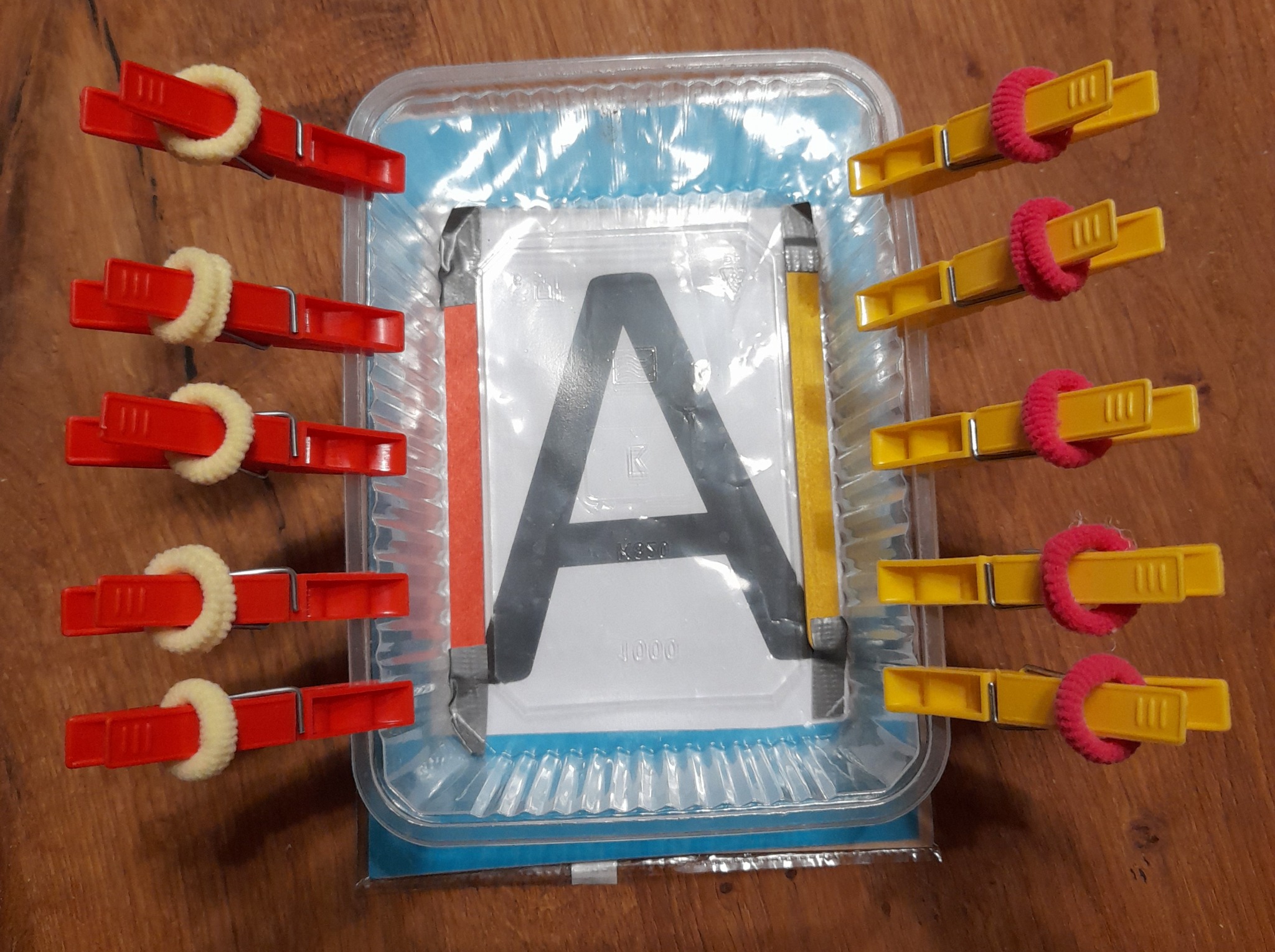 Szablon litery A w plastikowym pudełku, przytrzymywany z boków czerwonymi i żółtymi klamerkami z kolorowymi gumkami, na drewnianym podłożu.