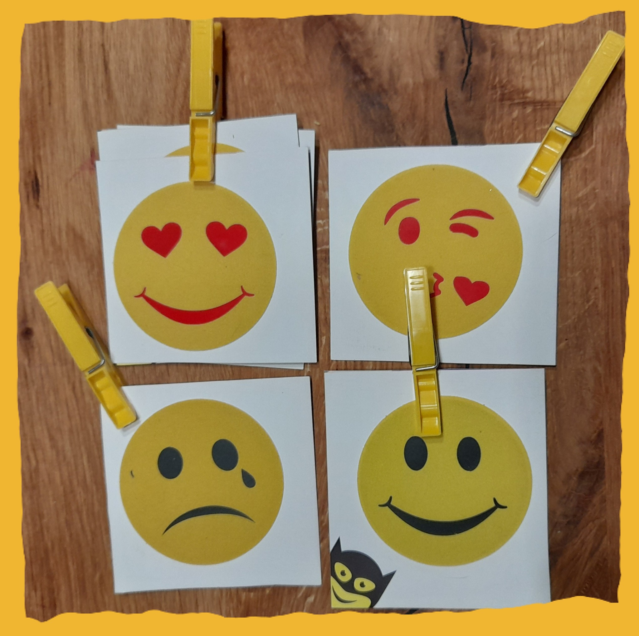 Cztery kartki z emotikonami przymocowane żółtymi klamerkami do drewnianej powierzchni: zakochana, puszczająca buziaka, smutna i uśmiechnięta, z małą naklejką nietoperza w rogu.