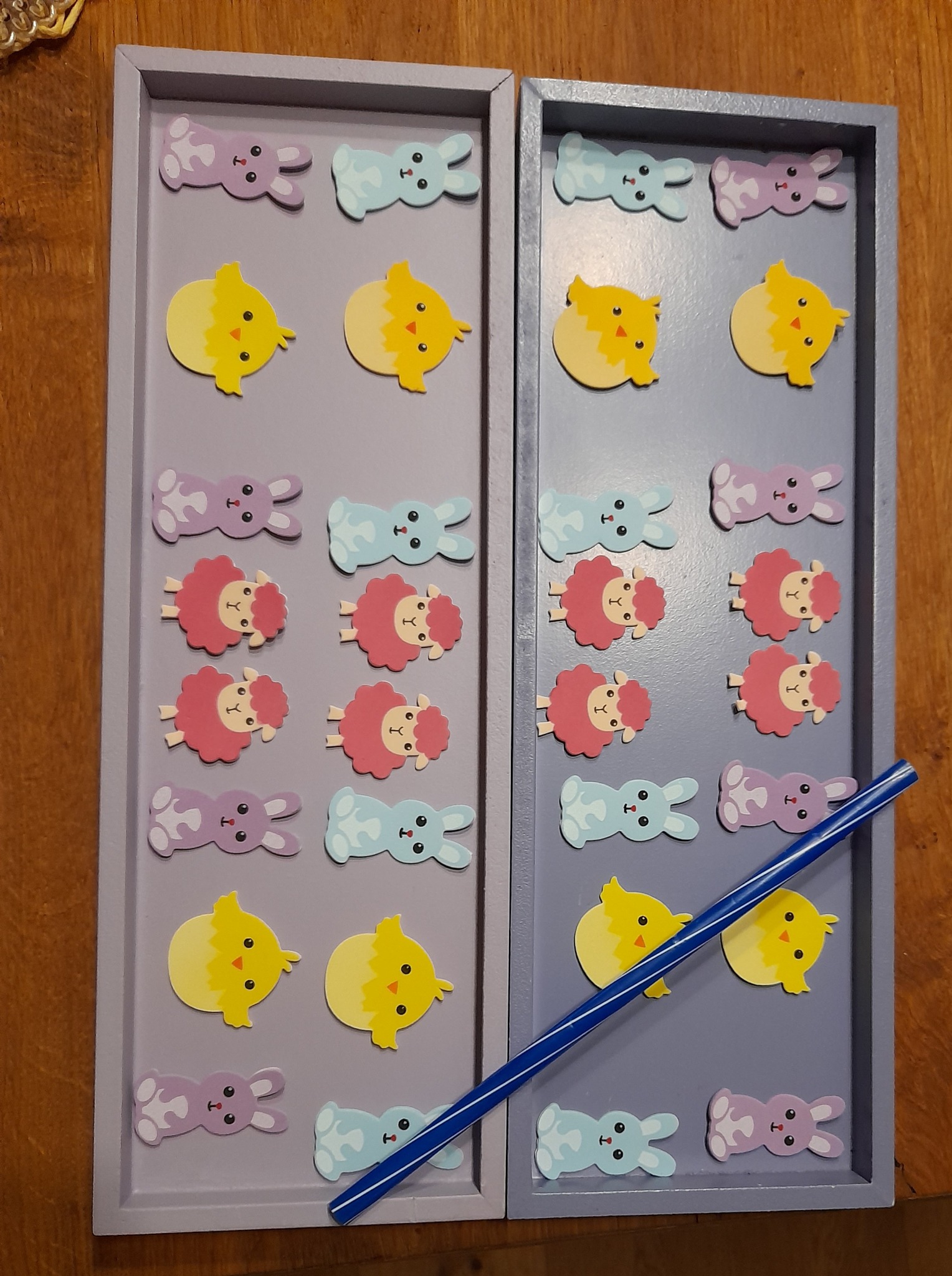 Dwie drewniane tace w odcieniach fioletu i szarości, ozdobione naklejkami z motywem wielkanocnym: króliczki, kurczaczki i owieczki, z niebieską słomką leżącą na jednej z tac.