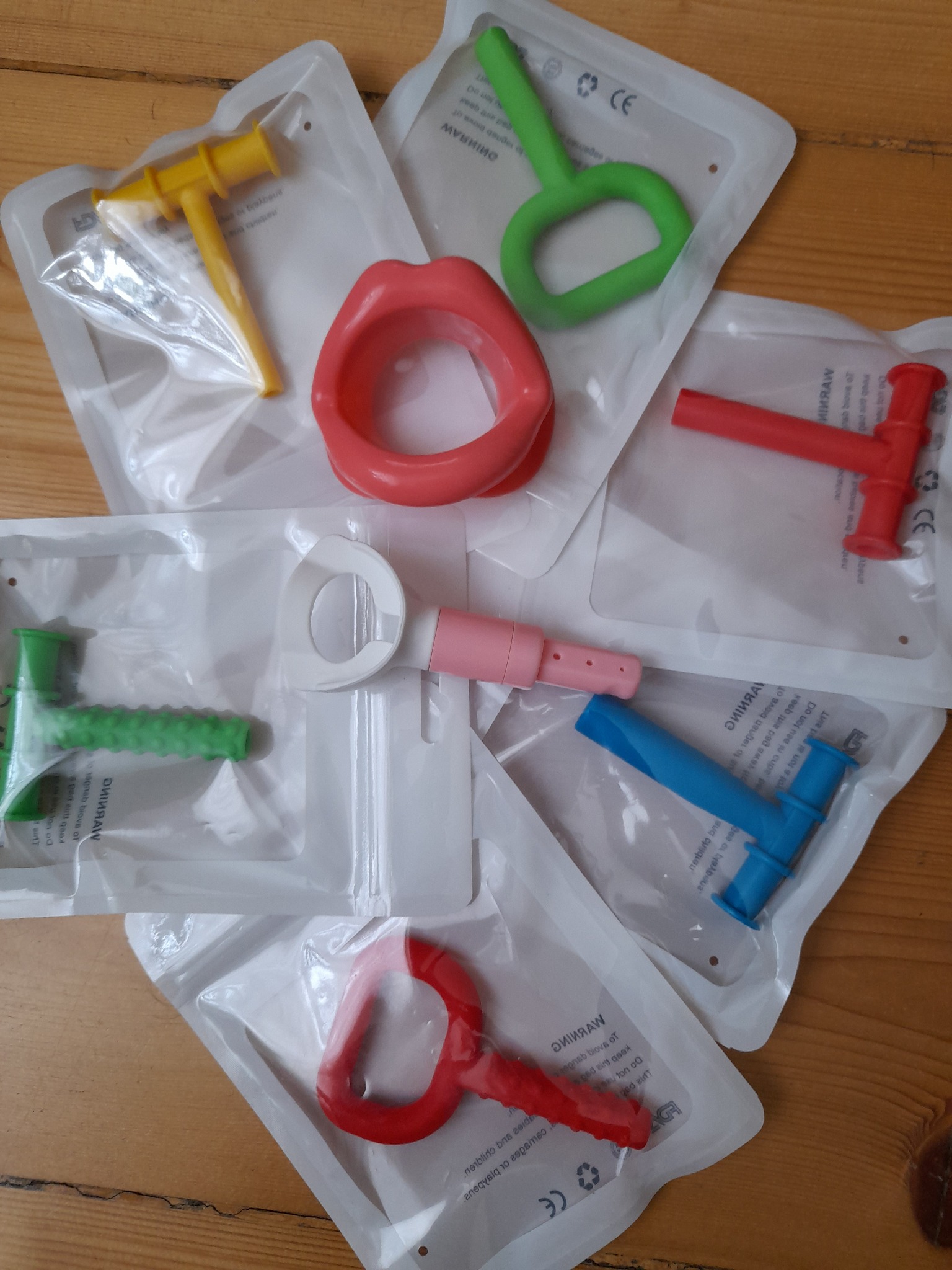 Kolorowe narzędzia do ćwiczeń logopedycznych, zapakowane w przezroczyste woreczki, leżą na drewnianym blacie.