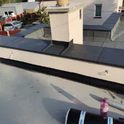 Wykończony dach z czarną papą, obróbki blacharskie w kolorze antracytowym, komin otynkowany na beżowo, widoczna rolka papy Soprema oraz butelka płynu Polaris.