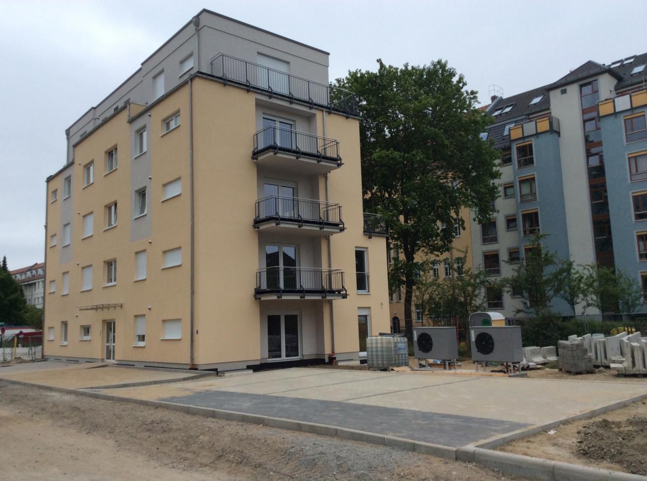 Nowo wybudowany budynek wielorodzinny z balkonami, widoczne elementy infrastruktury na zewnątrz, w tle inne budynki i drzewa.