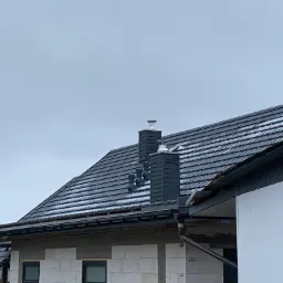 Nowy, ciemny dach z dachówki ceramicznej z kominem z wkładami kominowymi, częściowo pokryty śniegiem, na budynku w trakcie budowy z białych bloczków, pochmurne niebo w tle.