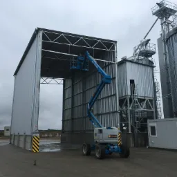 Budowa hali stalowej z użyciem podnośnika koszowego Genie Z-51/30J na tle silosów zbożowych i kontenera biurowego w pochmurny dzień.