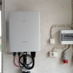 Biała skrzynka falownika Sungrow zamontowana na teksturowanej ścianie obok skrzynki bezpiecznikowej, gniazdka i kabli w peszlach.