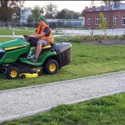 Pracownik w kamizelce odblaskowej kosi trawnik traktorkiem John Deere w parku miejskim, widoczny budynek w tle.