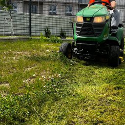 BIUREN Renata Pawlik - Zielony traktor kosi trawę na działce z ogrodzeniem z siatki. Część trawy skoszona, część jeszcze nie. Widoczne białe kwiaty na trawniku. Operator w kamizelce odblaskowej.