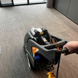 BIUREN Renata Pawlik - Szorowarka do dywanów Procarpet 30 w trakcie pracy na szarym dywanie w biurze, widoczna ręka operatora i fragment okna z widokiem na parking.