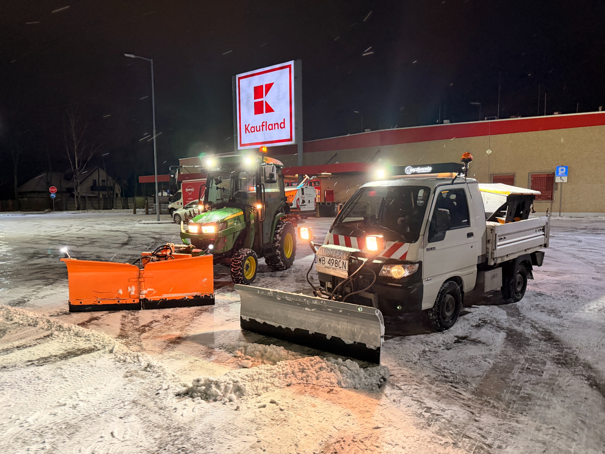 Dwa pojazdy do odśnieżania (ciągnik i mała ciężarówka) z pługami na parkingu Kauflandu, nocą, podczas opadów śniegu. Widoczne ślady opon na śniegu.