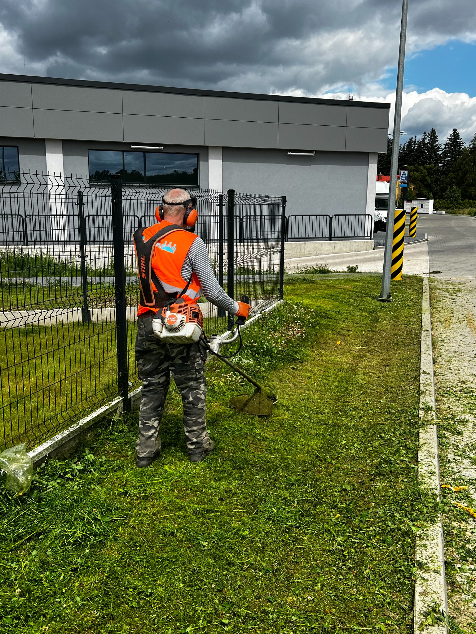 Mężczyzna w kamizelce tnący trawę przy ogrodzeniu, używając podkaszarki Stihl. Świeżo skoszona trawa leży na chodniku. Pochmurne niebo w tle.
