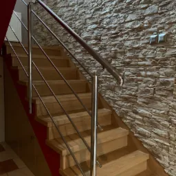 Drewniane schody z balustradą ze stali nierdzewnej, ściana wyłożona kamieniem dekoracyjnym, widoczne dwa włączniki światła.