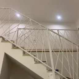 Biała balustrada schodowa z ukośnymi prętami, zabezpieczona taśmą malarską na stopniach podczas remontu, oświetlona dwoma punktowymi lampami sufitowymi.