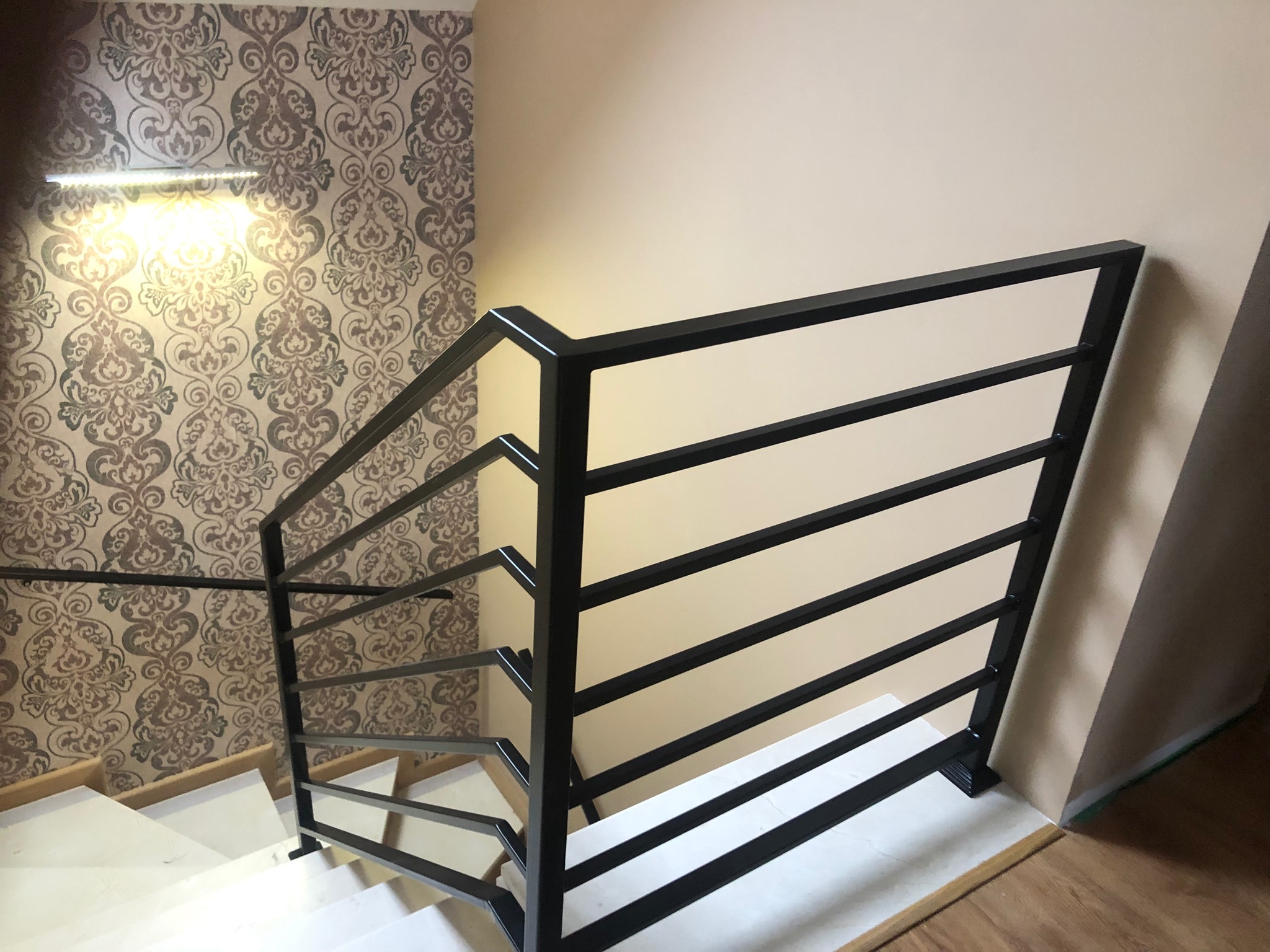 Nowoczesna, czarna balustrada schodowa z poziomych prętów, zamontowana przy białych schodach i ścianie z wzorzystą tapetą, oświetlona górnym światłem.