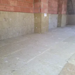 Ocieplenie poddasza wełną mineralną, widoczne mocowania z białymi kołkami, fragment ściany z cegły dziurawki, surowe drewno konstrukcyjne dachu