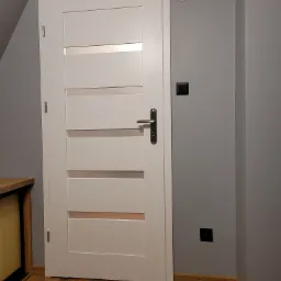 Białe drzwi wewnętrzne z poziomymi przeszkleniami, zamontowane w szarej ścianie, widoczne gniazdka elektryczne i fragment drewnianej podłogi.