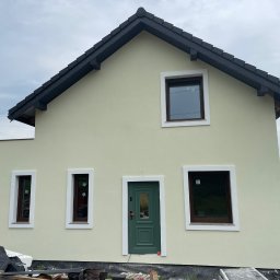 Mariusz Antonik - Dwukondygnacyjny dom z jasną elewacją, ciemną dachówką i zielonymi drzwiami wejściowymi, widok z zewnątrz na etapie wykończenia z widocznymi materiałami budowlanymi i rusztowaniem.