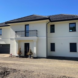 Mariusz Antonik - Dwukondygnacyjny dom z jasną elewacją, czarną dachówką i balkonem z kutą balustradą, otoczony niezagospodarowanym terenem.