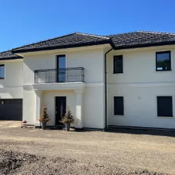 Dwukondygnacyjny dom z jasną elewacją, czarną dachówką i balkonem z kutą balustradą, otoczony niezagospodarowanym terenem.