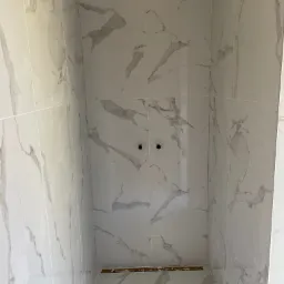 Wnętrze kabiny prysznicowej wyłożonej białymi płytkami imitującymi marmur, widoczne otwory na baterię i odpływ liniowy w podłodze, prace w trakcie.