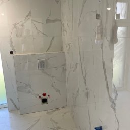 Mariusz Antonik - Wnętrze łazienki w trakcie wykańczania, widoczne białe płytki imitujące marmur na ścianach i podłodze, podtynkowa spłuczka WC z przyciskiem, wystające przewody instalacji wodnej i elektrycznej...