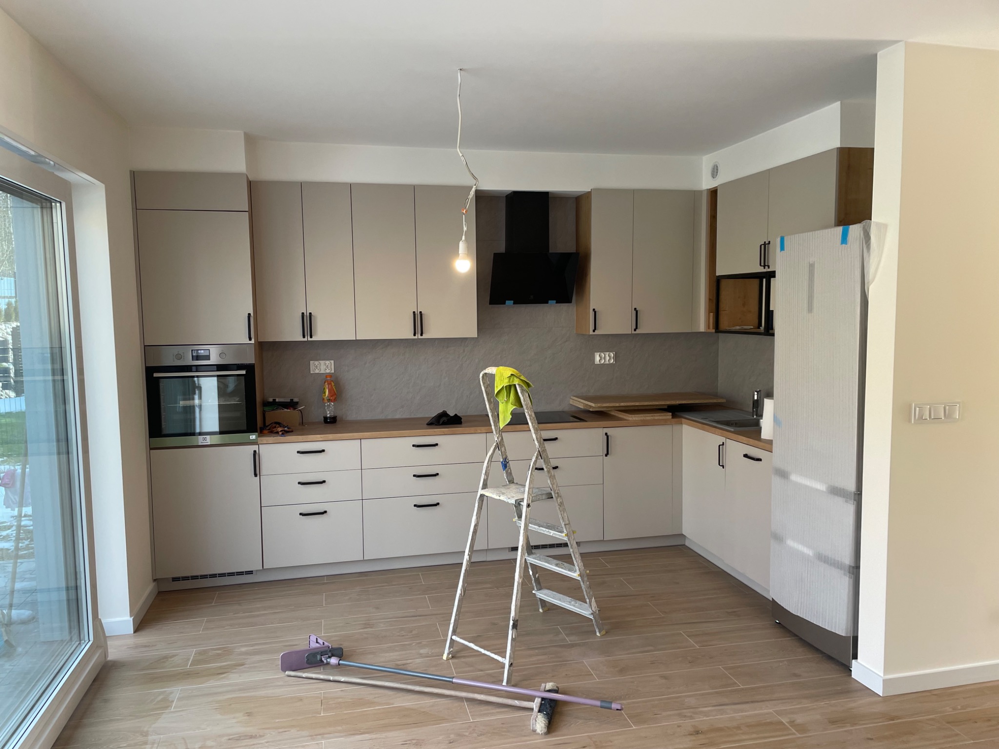 Jasna kuchnia w trakcie remontu: widoczna drabina malarska z zieloną ścierką, częściowo rozpakowana lodówka, drewnopodobne płytki podłogowe i surowa żarówka zwisająca z sufitu.