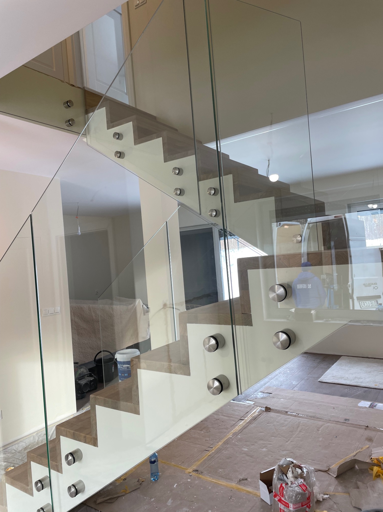 Szklana balustrada zamocowana punktowo do schodów z drewnianymi stopniami, widoczne zabezpieczenia podłogi kartonem podczas montażu.