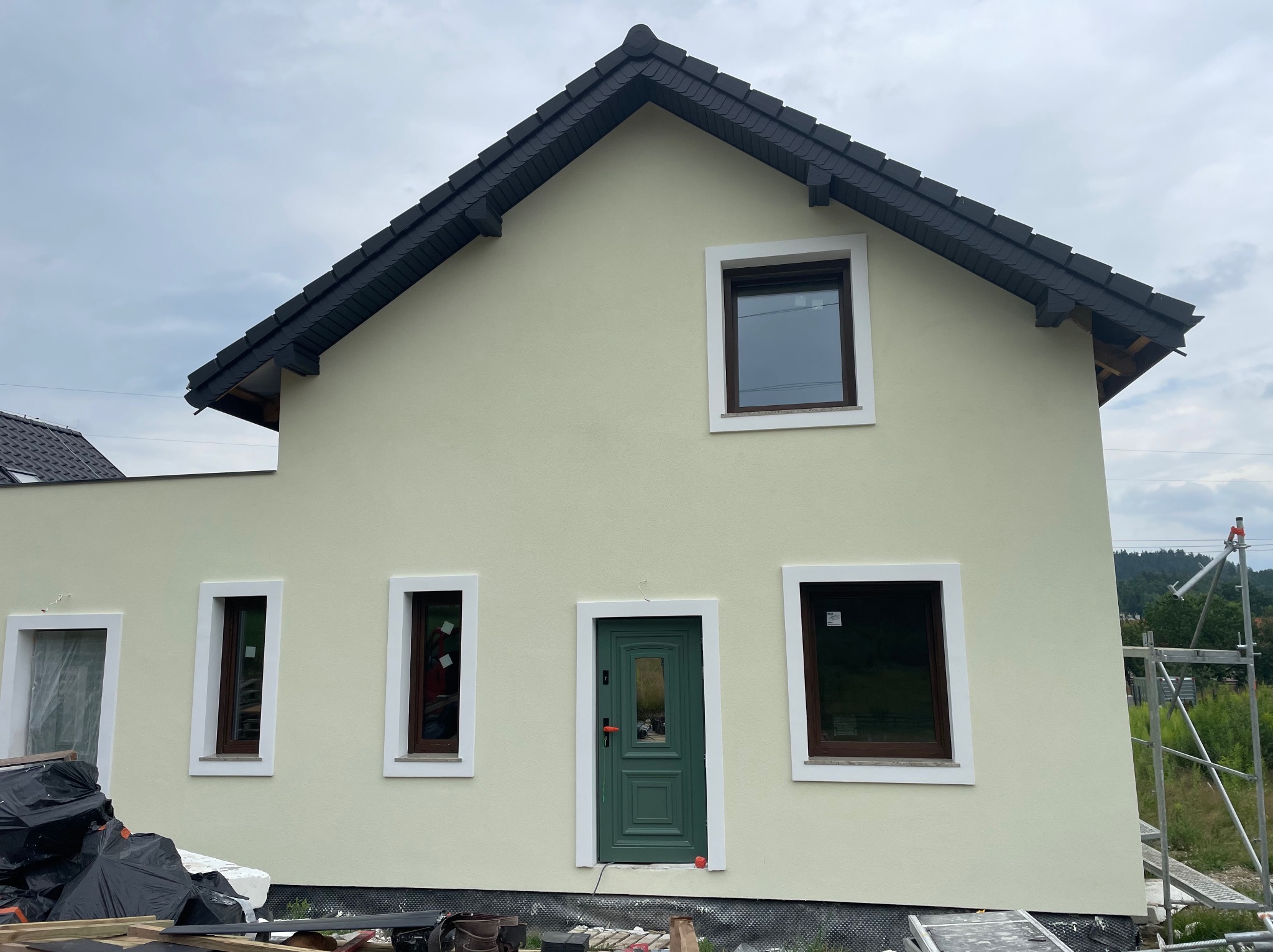 Dwukondygnacyjny dom z jasną elewacją, ciemną dachówką i zielonymi drzwiami wejściowymi, widok z zewnątrz na etapie wykończenia z widocznymi materiałami budowlanymi i rusztowaniem.