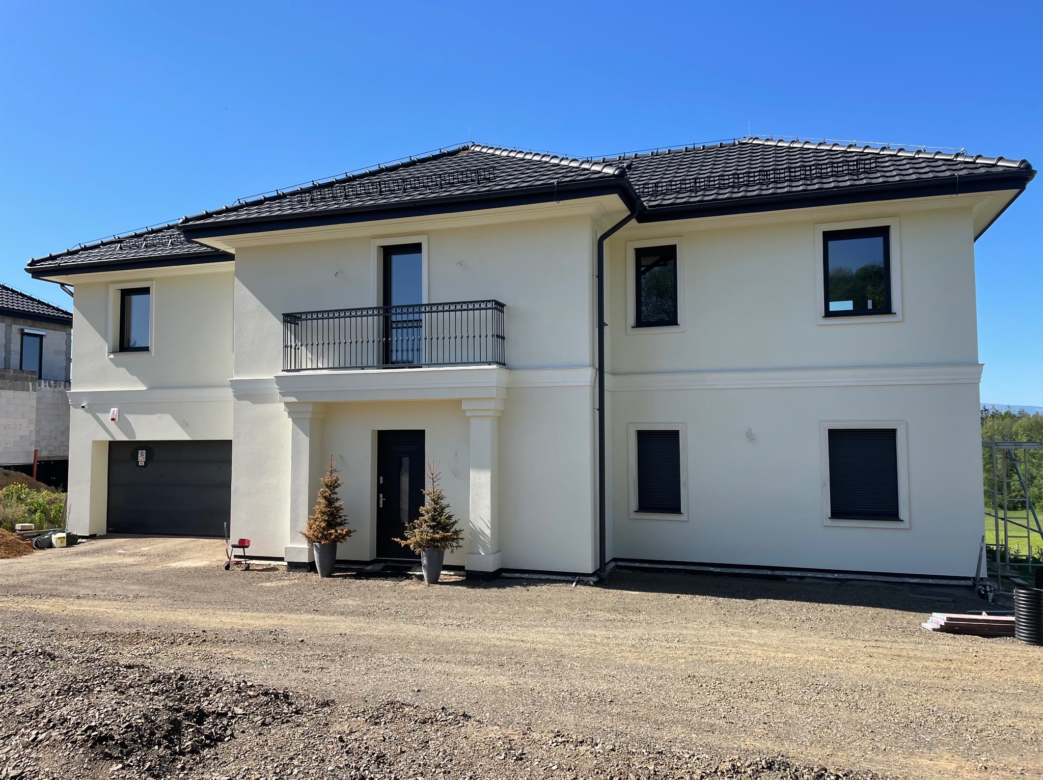 Dwukondygnacyjny dom z jasną elewacją, czarną dachówką i balkonem z kutą balustradą, otoczony niezagospodarowanym terenem.