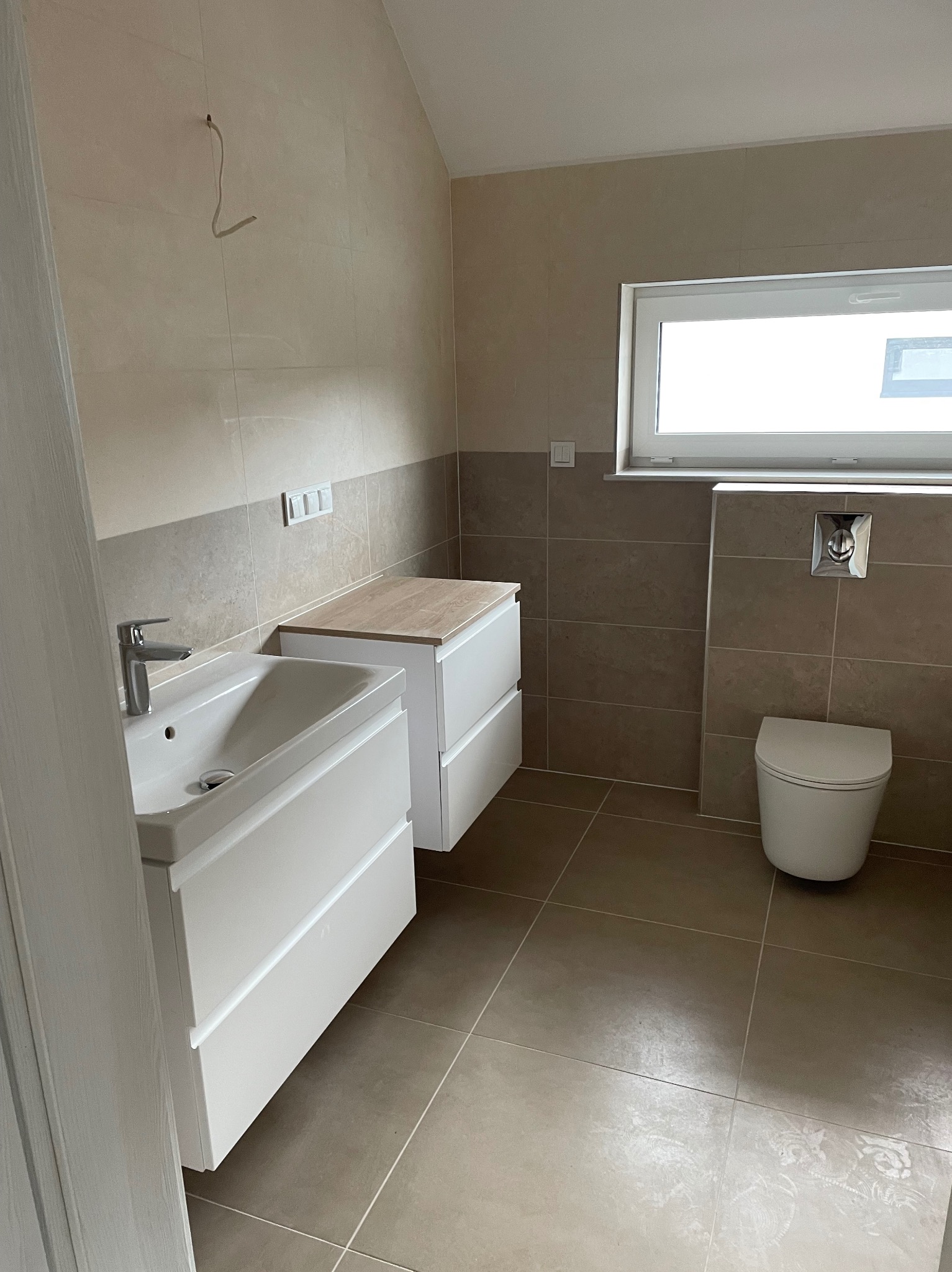 Nowoczesna łazienka z beżowymi płytkami, białą umywalką z szafką, toaletą podwieszaną i drewnianym blatem na szafce obok umywalki. Widoczny wystający kabel ze ściany.