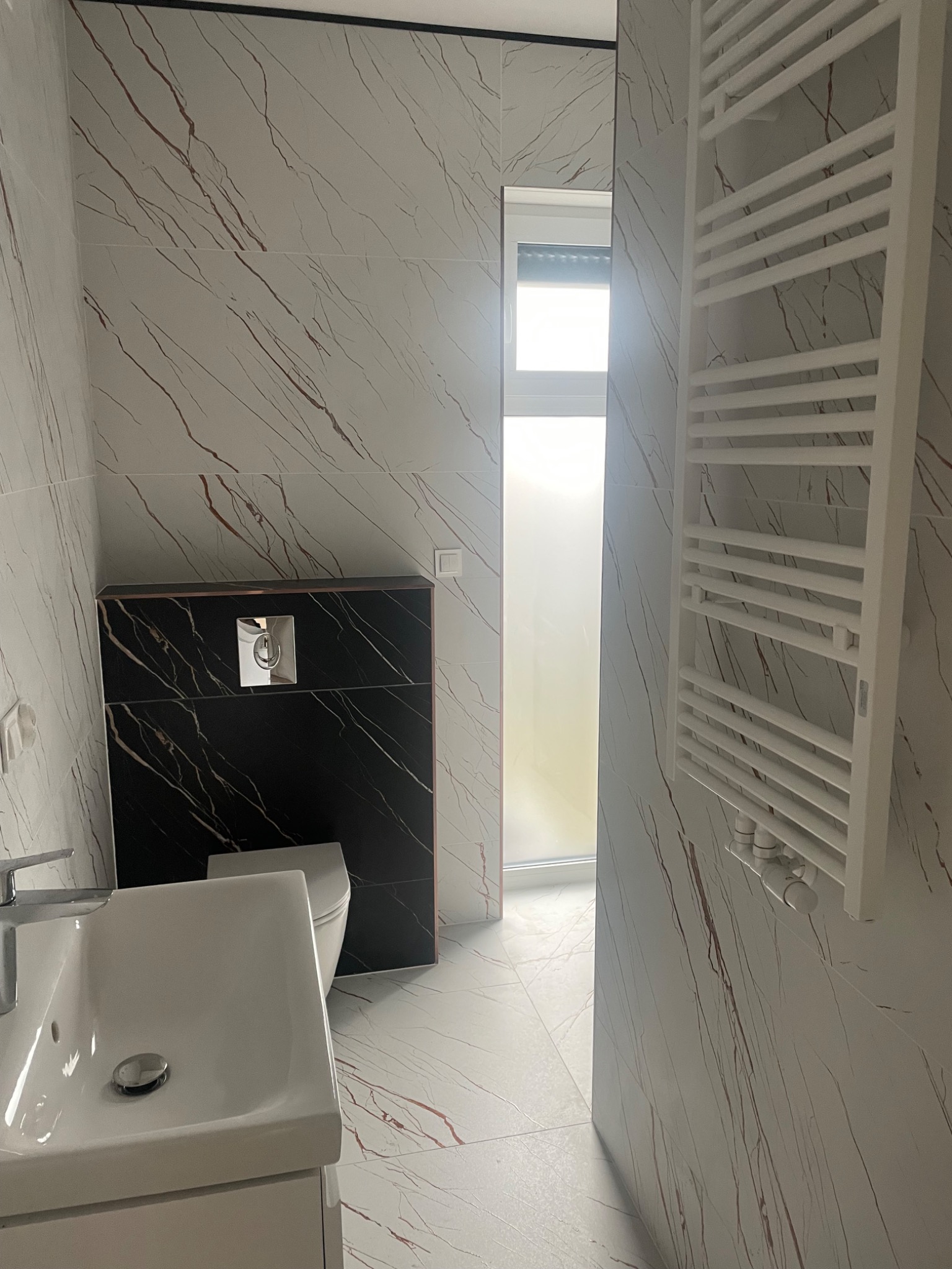 Wykończona łazienka w Jeleniej Górze z marmurowymi płytkami, wiszącą toaletą z czarną obudową i białym grzejnikiem drabinkowym.