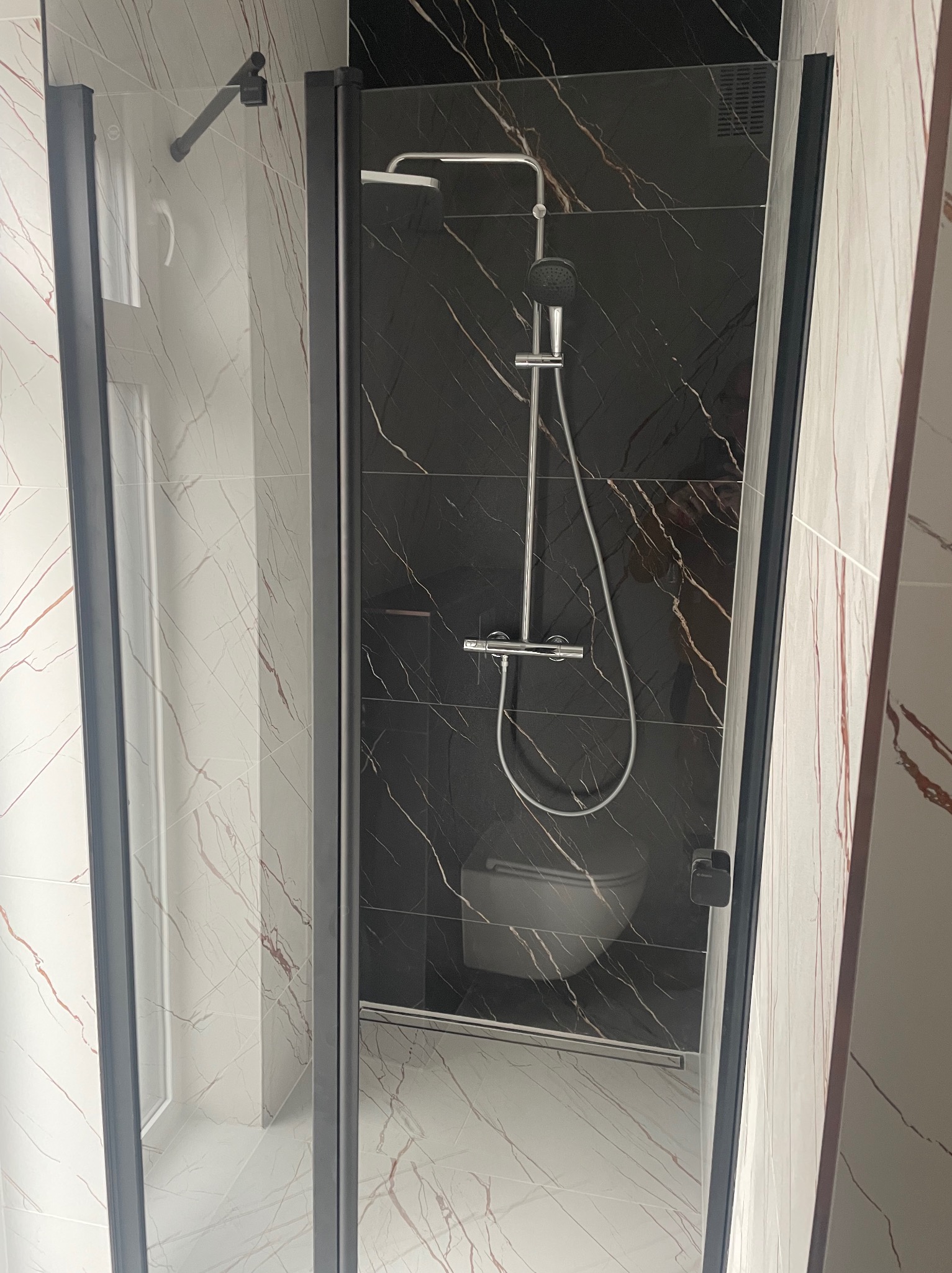 Nowoczesna kabina prysznicowa typu walk-in z czarnymi profilami, marmurowe płytki w odcieniach bieli i czerni z widoczną toaletą w tle.